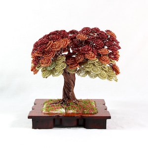 Könnte beinhalten: Eine Drahtskulptur eines Baumes mit einem verdrehten Stamm und einer belaubten Krone. Die Blätter sind aus Draht und in Rot-, Orange- und Grüntönen gefärbt. Der Baum steht auf einem Holzsockel.