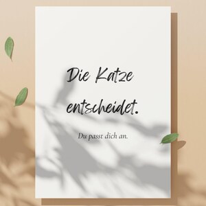 May include: A white rectangular print with the text "Die Katze entscheidet. Du passt dich an." in black script. The print is on a beige background with leaf shadows and three green leaves.