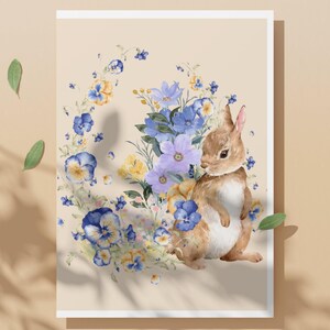 May include: A watercolor illustration of a brown bunny standing next to a floral wreath. The wreath features blue, yellow, and purple flowers. The background is a light beige color, and the image is framed in white.