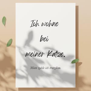 May include: A white rectangular card with black script that reads "Ich wohne bei meiner Katze. Miete zahle ich trotzdem." Green leaves and shadows are visible. The card is on a beige background.