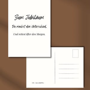 May include: Two white postcards on a brown background. The front postcard has black text that reads "Zum Jubil&auml;um, Du machst den Unterschied, Und rettest &ouml;fter den Morgen." The back postcard has lines for writing.