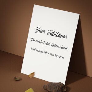 May include: A white card with black script that reads "Zum Jubil&auml;um, Du machst den Unterschied, Und rettest &ouml;fter den Morgen." The card is propped up on a brown surface with a few leaves and a rock.