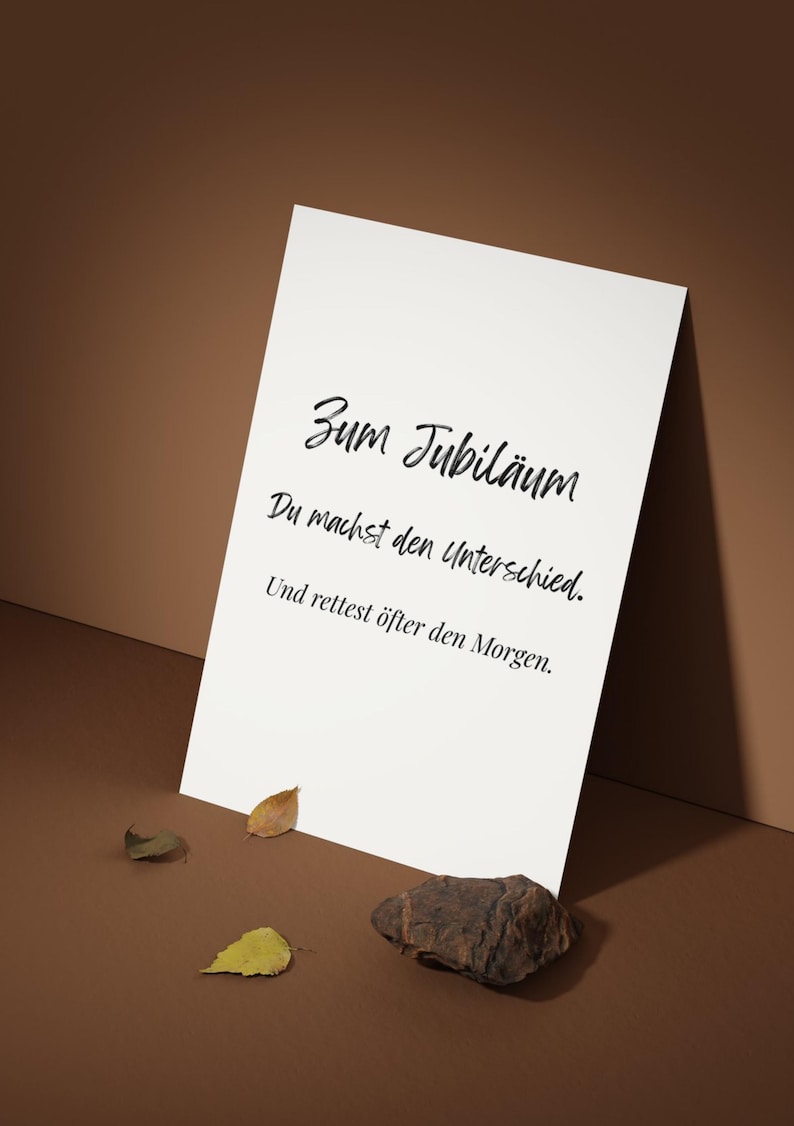 May include: A white card with black script that reads "Zum Jubil&auml;um, Du machst den Unterschied, Und rettest &ouml;fter den Morgen." The card is propped up on a brown surface with a few leaves and a rock.