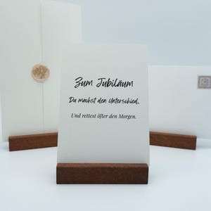May include: A white card with the text "Zum Jubil&auml;um" and other German text, displayed on a dark brown wooden stand. Two white envelopes are in the background, one with a gold seal.