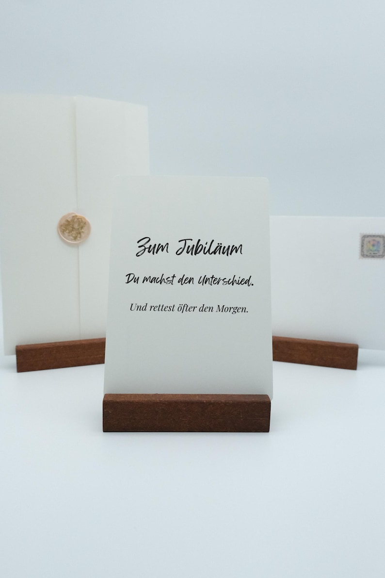 May include: A white card with the text "Zum Jubil&auml;um" and other German text, displayed on a dark brown wooden stand. Two white envelopes are in the background, one with a gold seal.