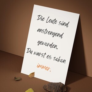 May include: A white card with black script that reads "Die Leute sind anstrengend geworden. Du warst es schon immer." The word "immer" is in orange. The card is on a brown surface with leaves and a rock.