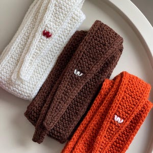 Può includere: Tre strisce di tessuto a maglia in bianco, marrone e arancione. Ogni striscia ha una piccola lettera "W" ricamata in un colore a contrasto.