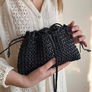 Könnte beinhalten: Eine schwarze gewebte Handtasche mit Kordelzugverschluss. Die Tasche wird von einer Person gehalten, die eine weiße Bluse mit Ösen trägt. Die Textur der Tasche scheint aus einem dicht gewebten Material zu bestehen, und der Kordelzug besteht aus einer passenden schwarzen Kordel.
