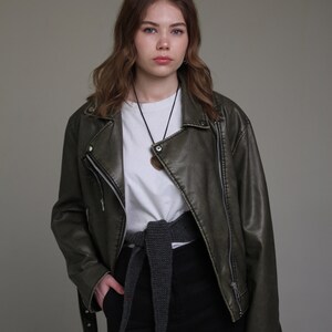 May include: A woman wearing a dark green leather jacket with a white t-shirt underneath. She is wearing a gray knitted belt and black pants.