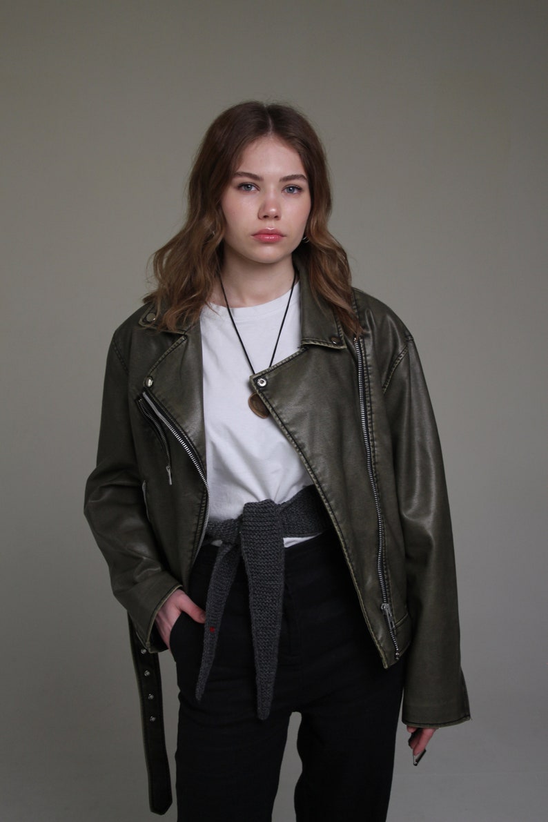 May include: A woman wearing a dark green leather jacket with a white t-shirt underneath. She is wearing a gray knitted belt and black pants.