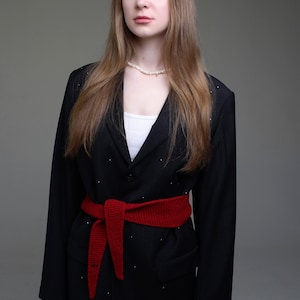May include: A woman wearing a black blazer with a red knitted belt and a pearl necklace. The blazer has a pinstripe pattern.
