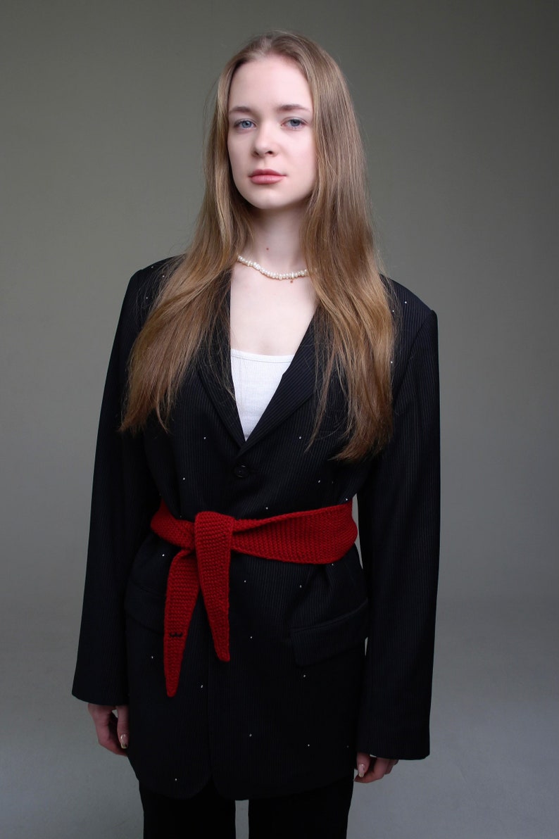 May include: A woman wearing a black blazer with a red knitted belt and a pearl necklace. The blazer has a pinstripe pattern.