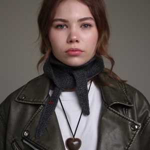 May include: A woman wearing a dark gray knitted scarf and a brown leather jacket with a zipper. She is wearing a white t-shirt and a brown heart-shaped pendant necklace.