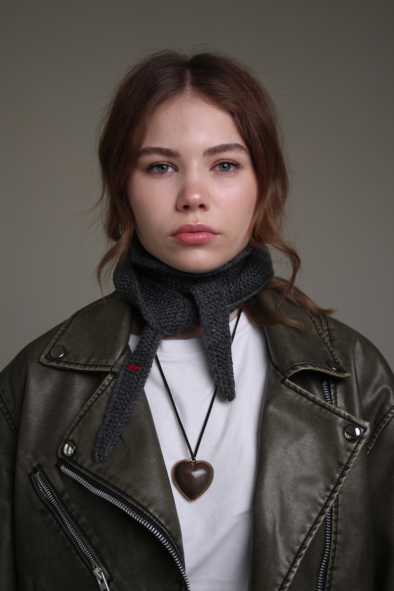 May include: A woman wearing a dark gray knitted scarf and a brown leather jacket with a zipper. She is wearing a white t-shirt and a brown heart-shaped pendant necklace.