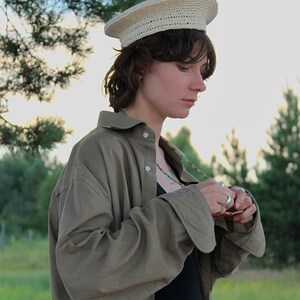 Peut inclure: Un chapeau en crochet beige clair avec un dessus plat et un large bord. Le chapeau est port&eacute; avec une chemise boutonn&eacute;e vert olive &agrave; manches longues et un body noir. Un collier de perles color&eacute;es est &eacute;galement visible.