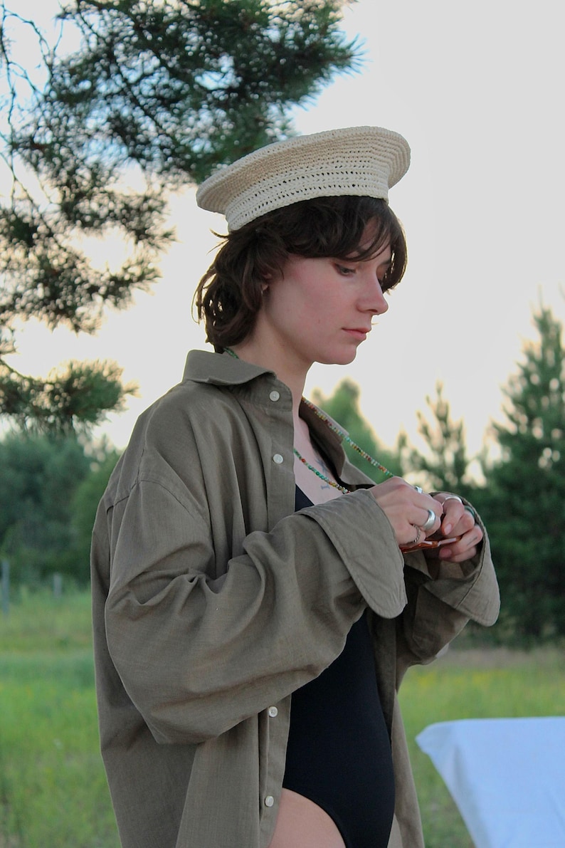 Peut inclure: Un chapeau en crochet beige clair avec un dessus plat et un large bord. Le chapeau est port&eacute; avec une chemise boutonn&eacute;e vert olive &agrave; manches longues et un body noir. Un collier de perles color&eacute;es est &eacute;galement visible.