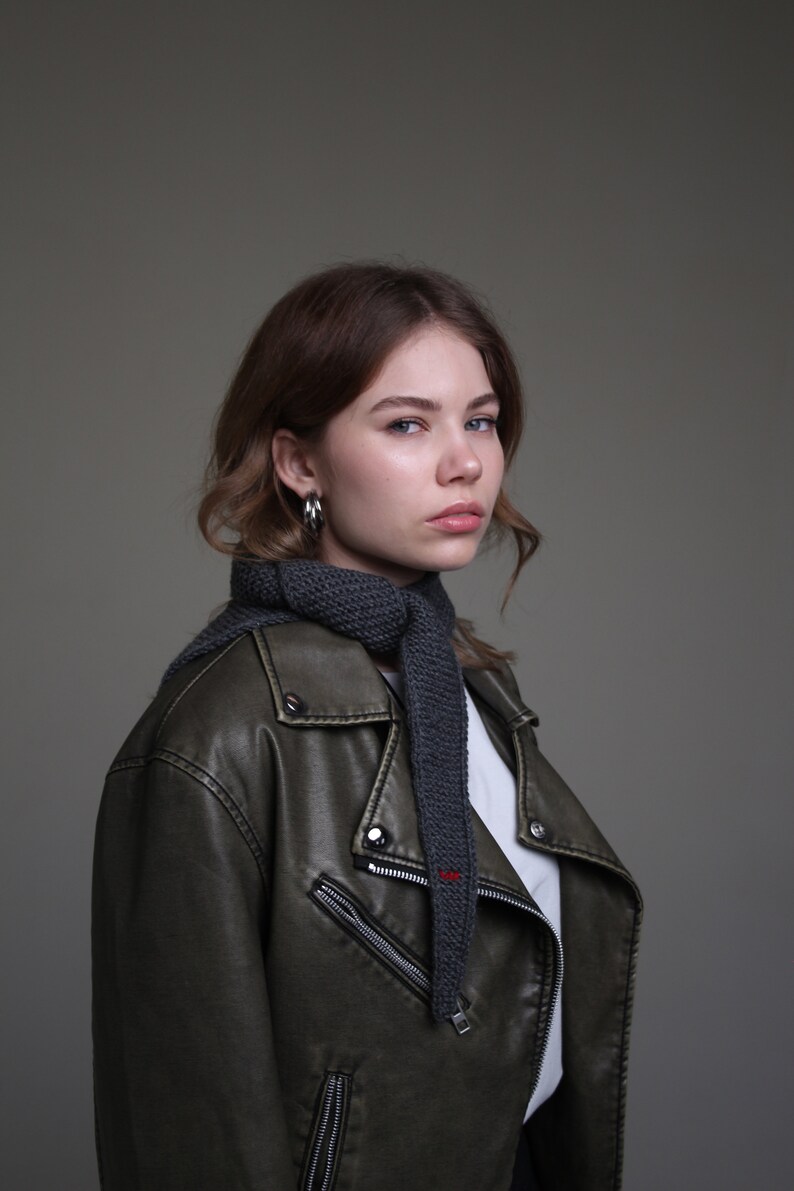May include: A woman with long brown hair wearing a dark green leather jacket with a gray knitted scarf.