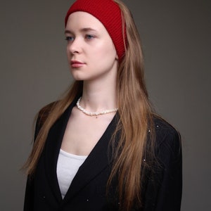 May include: A woman with long brown hair wearing a red headband and a black blazer with a white undershirt. She is wearing a pearl necklace.