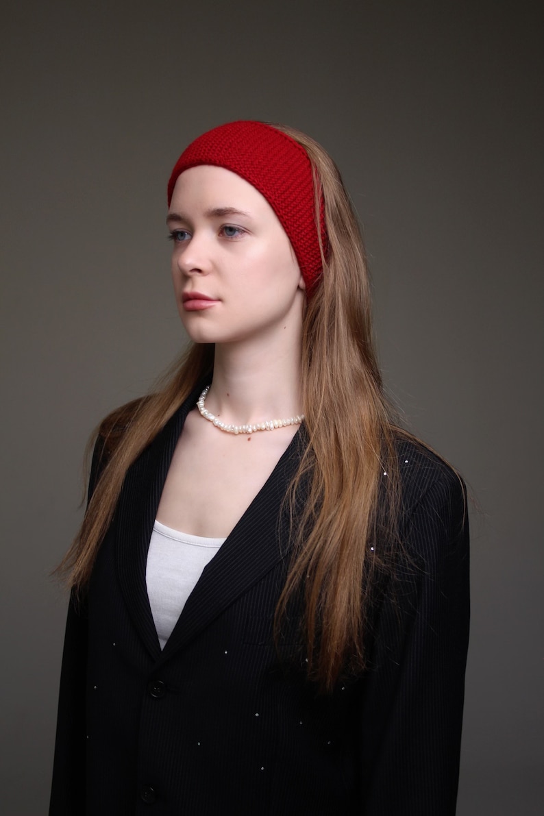 May include: A woman with long brown hair wearing a red headband and a black blazer with a white undershirt. She is wearing a pearl necklace.