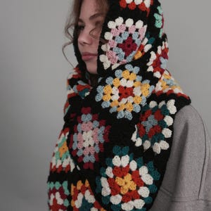 Puede incluir: Una bufanda de ganchillo negra con un patrón de cuadrados de la abuela de colores. La bufanda se lleva como capucha y presenta una variedad de colores, incluyendo rojo, amarillo, azul, verde y blanco.