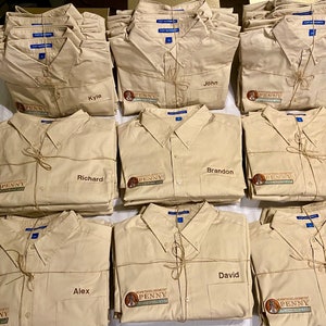 Puede incluir: Una pila de camisas de botones de color beige con un logotipo y los nombres John, Kyle, Ed, Richard, Joel, Brandon, David, Alex y Matthew bordados.