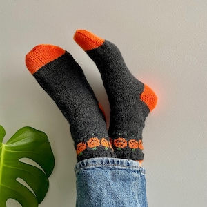 May include: A pair of black socks with orange tops and a pattern of orange pumpkins. The socks are being worn by someone wearing blue jeans.