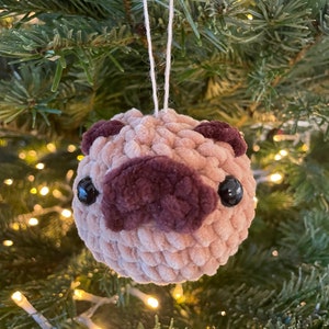Puede incluir: Un adorno de pug de crochet en beige y marrón, colgado de una cuerda blanca en un árbol de Navidad verde.