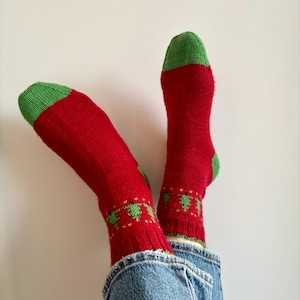 May include: A pair of red knit socks featuring green toes and heels, with a ribbed cuff and a small green tree pattern encircling the ankle area. The socks are worn with blue denim jeans, showcasing a classic knit texture and vibrant holiday-inspired color scheme.