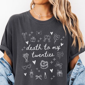 May include: A dark gray t-shirt with a white graphic design that says "death to my twenties" and features various celebratory icons like hearts, gifts, balloons, and champagne glasses. The word "PEPPER" is printed in white at the bottom of the shirt.