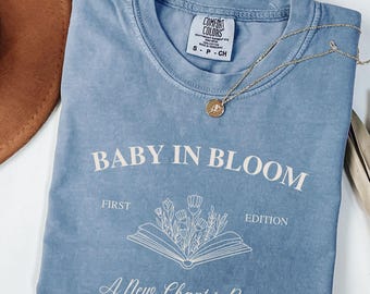 May include: A light blue t-shirt with a white graphic design that says "Baby in Bloom, First Edition, A New Chapter Begins, Published 2025". The graphic features a book with flowers growing out of it.