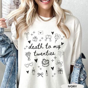 May include: White t-shirt with a black graphic that says "death to my twenties" surrounded by small black illustrations of hearts, gifts, balloons, glasses, lipstick, and other celebratory items. The text "IVORY" is in the bottom right corner.