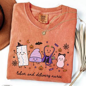 May include: A rust-colored t-shirt featuring a graphic of a cartoon fetal monitor strip, a purple beanie, a stethoscope, and a pink character. The text "labor and delivery nurse" is printed below. The shirt has a Comfort Colors brand label.