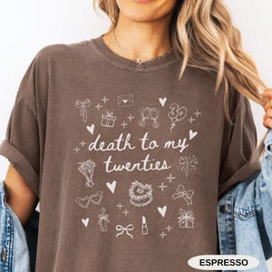 May include: A brown t-shirt with a white graphic that says "death to my twenties" and is surrounded by various white icons, including a gift box, a champagne flute, a lipstick, a bow, and a cake. The text "ESPRESSO" is printed in white at the bottom of the shirt.