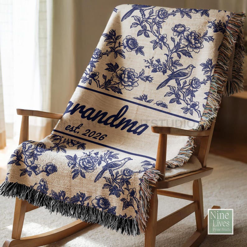 K&ouml;nnte beinhalten: Eine beige Decke mit blauem Blumen- und Vogelmuster liegt &uuml;ber einem Schaukelstuhl aus Holz. Auf der Decke stehen die Worte "grandma" und "est. 2026" in Blau. Die Decke hat einen Fransenbesatz.
