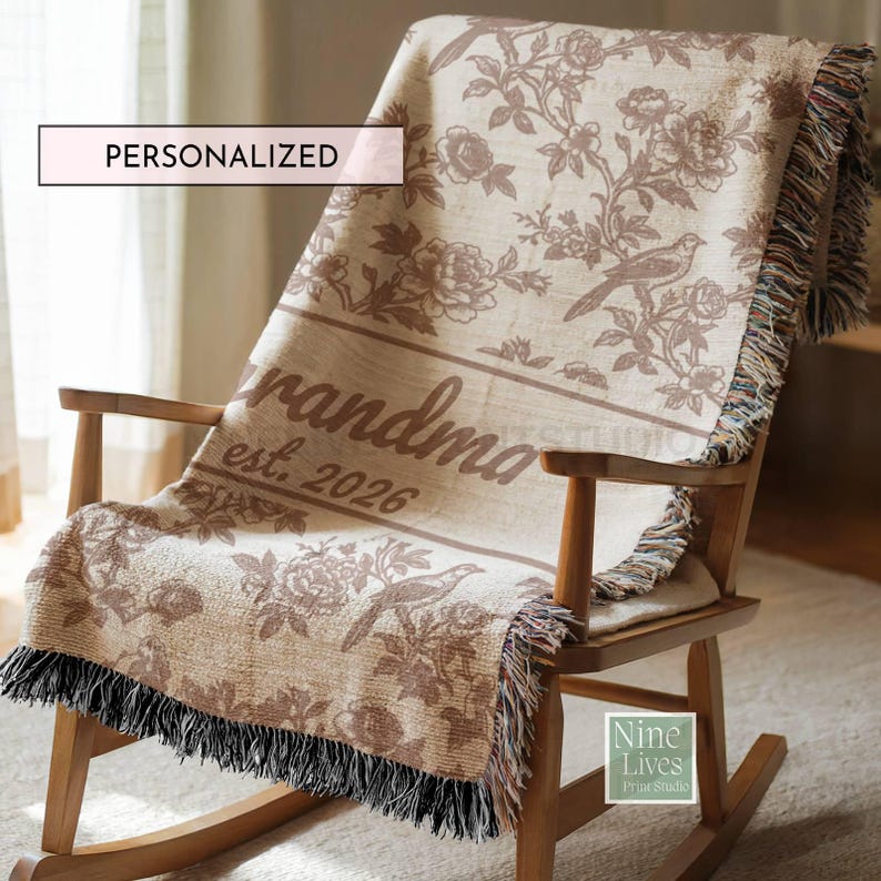 K&ouml;nnte beinhalten: Eine personalisierte Decke in Beige und Braun, die &uuml;ber einem Schaukelstuhl aus Holz drapiert ist. Die Decke hat ein Blumen- und Vogelmuster mit dem Wort &bdquo;Oma&ldquo; und dem Jahr &bdquo;est. 2026&ldquo; aufgedruckt. Die Decke hat einen Fransenbesatz.