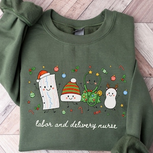 Könnte beinhalten: Ein olivgrünes Sweatshirt mit Rundhalsausschnitt und einer Grafik aus vier Zeichentrickfiguren: ein Herzmonitorstreifen mit Weihnachtsmütze, eine gestreifte Mütze, ein Weihnachtsbaum mit Stethoskop und ein Rentier. Text: labor and delivery nurse.