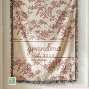 K&ouml;nnte beinhalten: Cremefarbene Decke mit einem sich wiederholenden Muster aus V&ouml;geln und floralen Elementen in Rosa. Das Wort "grandma" und das Jahr "2026" sind in der Mitte aufgedruckt. Die Decke hat einen Fransenrand und h&auml;ngt an einer Wand.