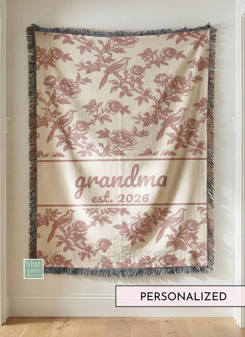 K&ouml;nnte beinhalten: Cremefarbene Decke mit einem sich wiederholenden Muster aus V&ouml;geln und floralen Elementen in Rosa. Das Wort "grandma" und das Jahr "2026" sind in der Mitte aufgedruckt. Die Decke hat einen Fransenrand und h&auml;ngt an einer Wand.