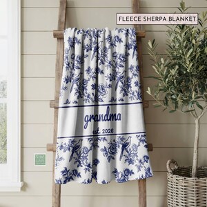 K&ouml;nnte beinhalten: Wei&szlig;e Fleece-Sherpa-Decke mit blauem Blumen- und Vogelmuster. Das Wort "grandma" und das Jahr "2026" sind blau gedruckt. Die Decke ist &uuml;ber eine Holzleiter drapiert.