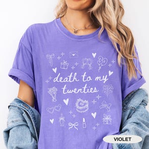 May include: A purple t-shirt with the text "death to my twenties" and white illustrations of party decorations, including a cake, balloons, and champagne glasses.