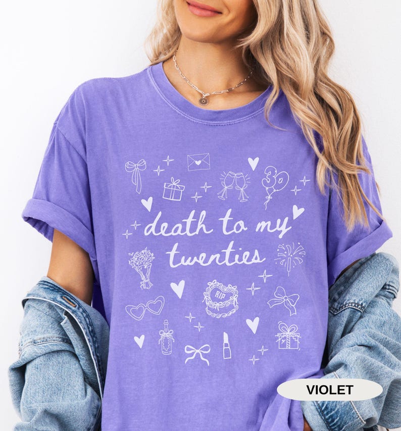 May include: A purple t-shirt with the text "death to my twenties" and white illustrations of party decorations, including a cake, balloons, and champagne glasses.