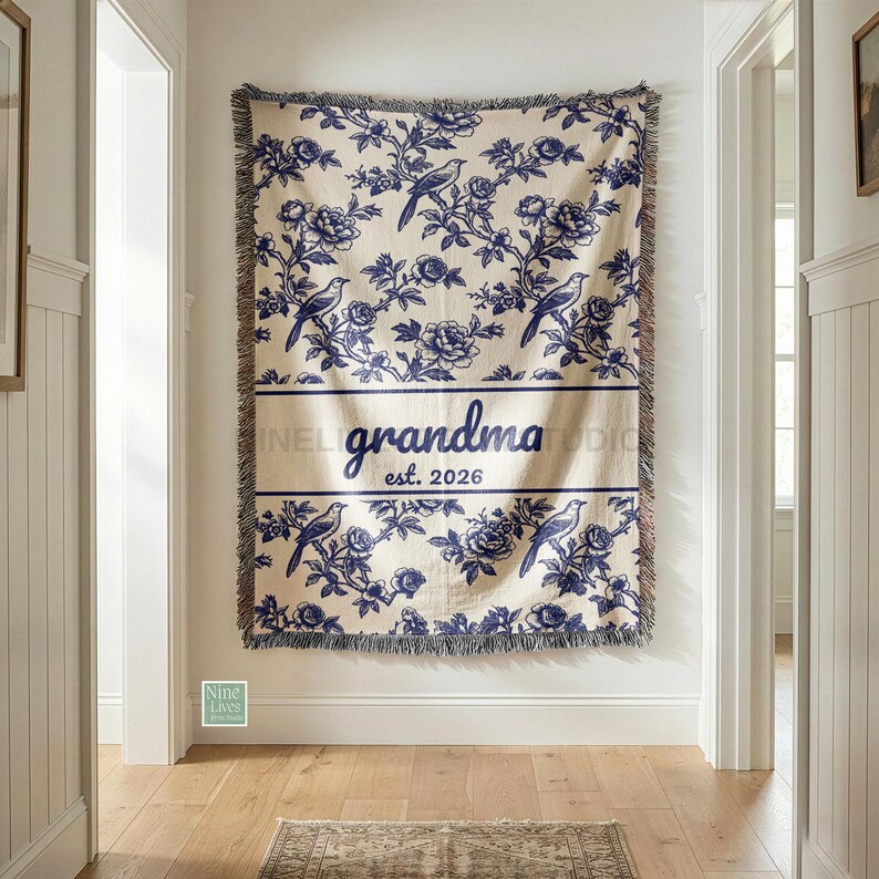 K&ouml;nnte beinhalten: Eine cremefarbene Decke mit blauem Blumen- und Vogelmuster. Das Wort "grandma" wird in einem Banner mit "est. 2026" darunter angezeigt. Die Decke hat Fransen an den R&auml;ndern und h&auml;ngt an einer wei&szlig;en Wand.
