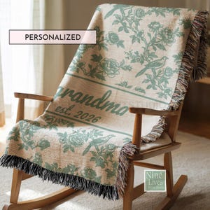 K&ouml;nnte beinhalten: Eine personalisierte beige Decke mit gr&uuml;nem Blumen- und Vogelmuster, drapiert &uuml;ber einem h&ouml;lzernen Schaukelstuhl. Auf der Decke steht "grandma est. 2026". Die Decke hat einen Fransenbesatz und das Wort "PERSONALISIERT" steht in einem wei&szlig;en Feld.