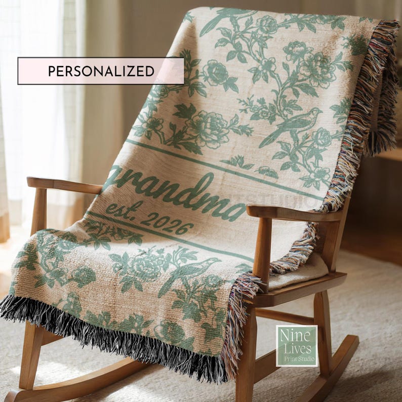 K&ouml;nnte beinhalten: Eine personalisierte beige Decke mit gr&uuml;nem Blumen- und Vogelmuster, drapiert &uuml;ber einem h&ouml;lzernen Schaukelstuhl. Auf der Decke steht "grandma est. 2026". Die Decke hat einen Fransenbesatz und das Wort "PERSONALISIERT" steht in einem wei&szlig;en Feld.