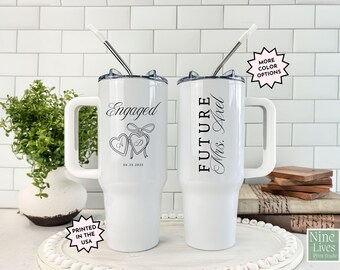 May include: Two white travel mugs with black text and a handle. One mug says "Engaged" with a heart design and date. The other says "FUTURE Mrs. Axel". Each mug has a lid and a straw. The mugs are printed in the USA.
