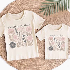Könnte beinhalten: Zwei cremefarbene T-Shirts mit floralen Designs. Eines der Shirts trägt den Schriftzug "mama" und das andere "mini" in Schreibschrift. Die floralen Designs umfassen rosa, graue und beige Blumen und Blätter. Die Shirts werden auf einer gewebten Matte präsentiert.