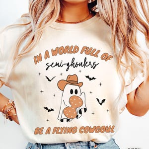 Könnte beinhalten: Elfenbeinfarbenes T-Shirt mit dem Text "IN A WORLD FULL OF semi-ghouls BE A FLYING COWGOUL". Die Grafik zeigt einen Geist mit Cowboyhut und Hufeisen. Schwarze Fledermäuse und Kreuze sind verstreut.
