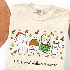 Könnte beinhalten: Ein cremefarbenes T-Shirt mit einem Comfort Colors-Etikett, das eine Grafik von vier Zeichentrickfiguren mit weihnachtlichem Zubehör zeigt, darunter ein Herzmonitorstreifen, eine Mütze, ein Baum mit Stethoskop und ein Rentier, mit dem Text labor and delivery nurse.