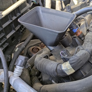 May include: A black plastic funnel is positioned over an engine's components, including hoses and wires. The engine bay is filled with various parts, with a focus on the funnel and surrounding elements. The image is taken from a close-up perspective.