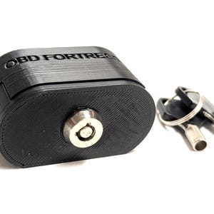 以下が含まれることがあります： 黒色のOBD FORTRESSセキュリティデバイス。キーロックと2つのキーが付いています。デバイスは楕円形で、上部に「OBD FORTRESS」の文字があります。キーは銀色です。
